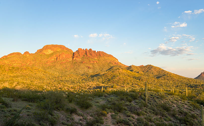 Setting sun casting a reddish-orange glow on Vulture Mountain, set against a robin blue sky with cumulus clouds and a shaded Sonoran Desert landscape.