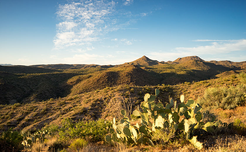 Sweeping view of the hills around Bagdad, Arizona, with a solitary prickly pear cactus in the foreground under a blue, partially cloudy sky.
