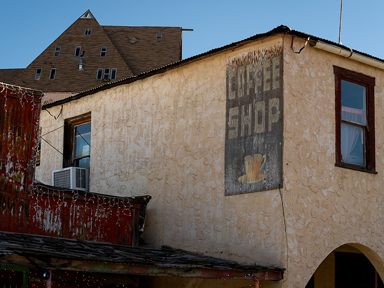 Weathered coffee shop sign on the side of a historic hotel in Oatman, Arizona, with an A-frame house looming in the background and a saloon to the left.