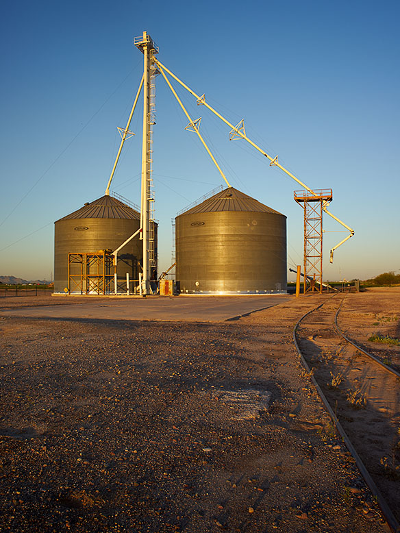 Two large grain storage silos standing beside the railroad tracks in Buckeye, Arizona, captured in the warm light of sunrise.