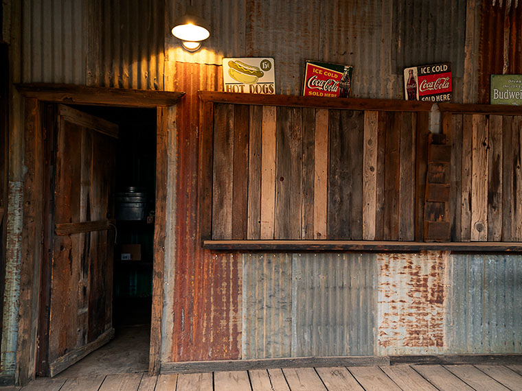 Weathered metal exterior of Vulture City's Cantina featuring vintage food and drink signs, captured in dim light after sunset.