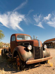 A rusted, vintage Ford truck stands in a ghost town, framed by timeworn buildings and a sky punctuated by twisted arrowhead-shaped cirrus clouds.