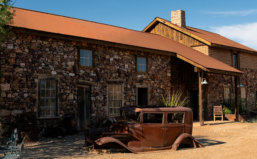 Historic rock-faced Assay Office in Vulture City with a rusted 30s-era Ford sedan in the foreground, set against a clear blue sky.