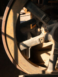Rusted drive wheel of Vulture City mine's headframe, with sunlit highlights against a shadowed background.