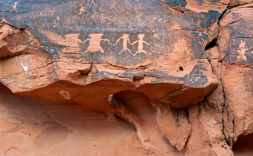 A detailed petroglyph panel featuring historical figures and animals, carved into desert varnish on red sandstone at Valley of Fire State Park, Nevada.