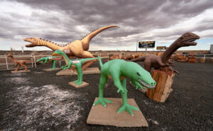 Wide-angle photo of playful dinosaur sculptures in a rock shop yard in Holbrook, Arizona, with petrified wood for sale