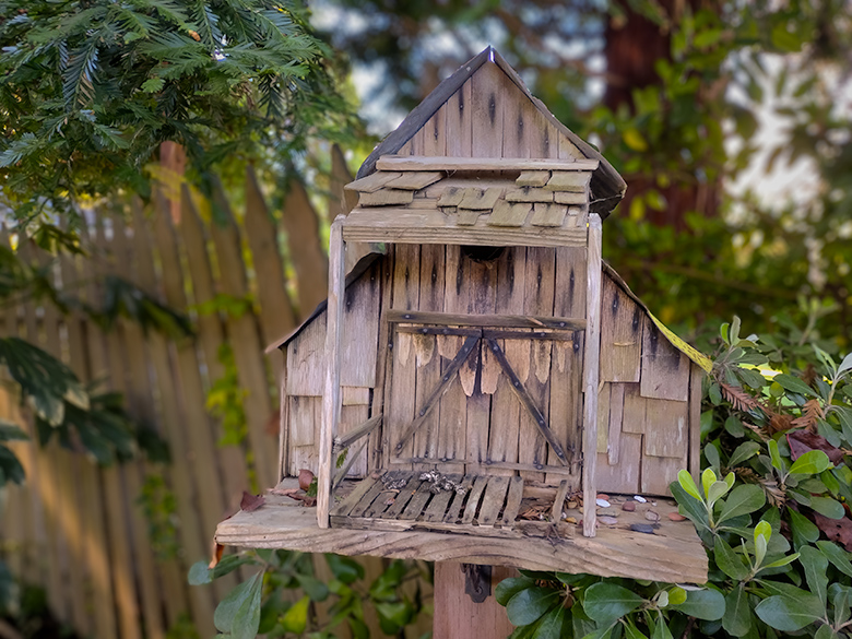 Old wooden barn-shaped birdhouse with broken boards, nestled among garden plants at Cambria Pines Lodge.