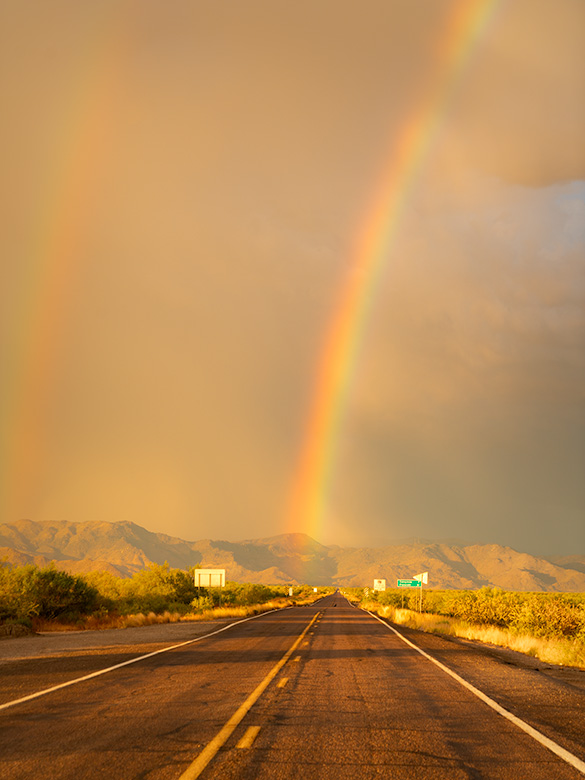 Double rainbows arching over State Route 71 with dark golden clouds and the Weaver Mountains in the background, Congress, Arizona.