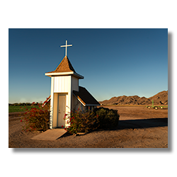 A quaint white chapel with a cross stands solitary among the lettuce fields under the expansive Arizona sky, inviting passersby for a moment of prayer and solitude.