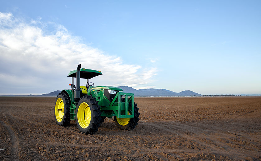 A new, green tractor with bright yellow wheels in a freshly plowed cotton field in Aguila, Arizona, with mountains in the distance.